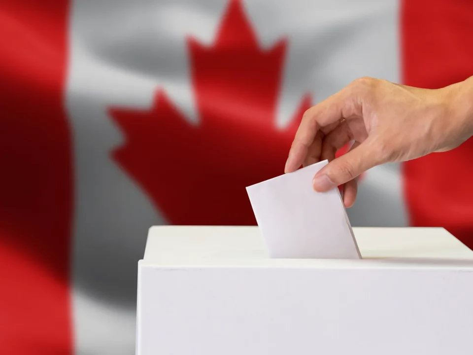 A voter's hand placing a paper ballot into a white ballot box, with a Canadian flag in the background.
