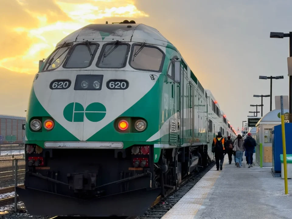 A green and white GO train at the Durham College Oshawa GO Station platform under a golden sky, with passengers walking along the platform